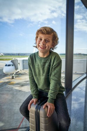 Foto de Encantado niño con el pelo rubio en traje casual sonriendo a la cámara mientras está sentado en la maleta cerca de la ventana y esperando la salida en el aeropuerto moderno - Imagen libre de derechos