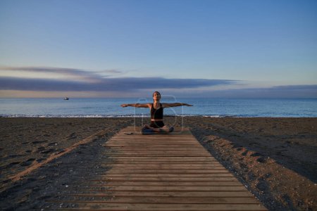 Téléchargez les photos : Femme paisible éloignée assise dans la posture Padmasana avec les bras écartés pendant la séance de yoga sur la promenade près de la mer le soir - en image libre de droit