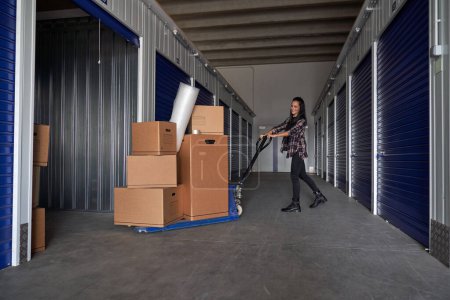 Photo for Full body side view of optimistic female using special manual pallet jack to roll packing boxes into rented self storage during moving day - Royalty Free Image