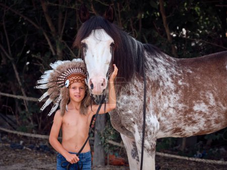 Positiver Frühchen-Junge in traditioneller nordamerikanischer Kopfbedeckung streichelt gehorsames Roan Horse mit Zügel und Zaumzeug und blickt in die Kamera, während er in der Nähe von Zaun und Bäumen auf dem Land steht
