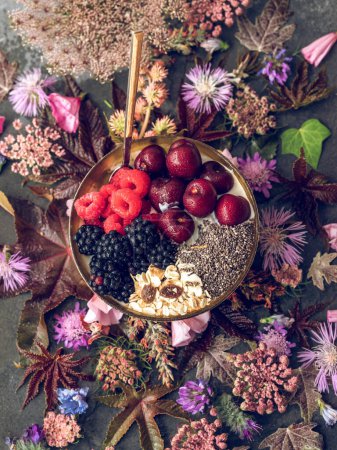 Photo for Top view of bowl filled with healthy breakfast topped with berries and chia seeds placed on table with flowers - Royalty Free Image