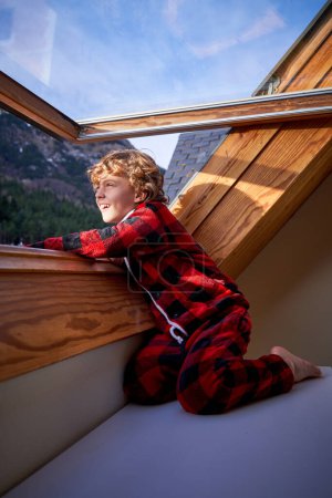 Téléchargez les photos : Vue latérale d'un gamin blond joyeux en pyjama rouge à carreaux chaud assis près de la fenêtre ouverte et regardant vers l'extérieur en observant la vue sur les montagnes et la forêt de l'hôtel - en image libre de droit
