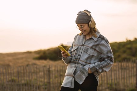 Photo for Self assured trendy female teenager in checkered shirt and hat smiling and listening to music in headphones while messaging on smartphone standing in rural field under cloudy sunset sky - Royalty Free Image