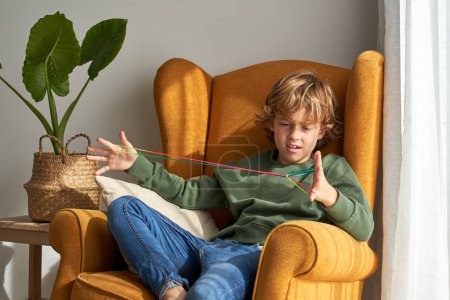 Téléchargez les photos : Enfant confus dans des vêtements décontractés assis dans un fauteuil ocre tout en jouant avec du fil de doigt arc-en-ciel et en faisant des figures de ficelle pendant le week-end à la maison - en image libre de droit