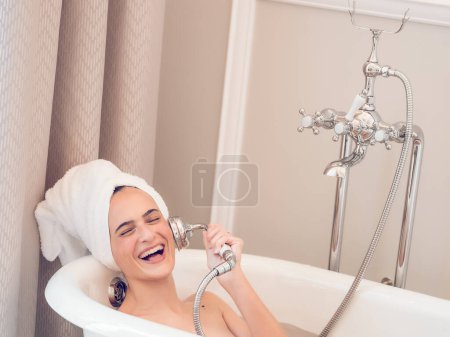 Photo for Delighted female with towel on head lying in soaker bathtub and imitating conversation on phone call with shower water can - Royalty Free Image