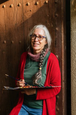 Foto de Encantada artista femenina senior con paleta de pintura y pinceles de pie cerca de la puerta de madera y mirando a la cámara - Imagen libre de derechos