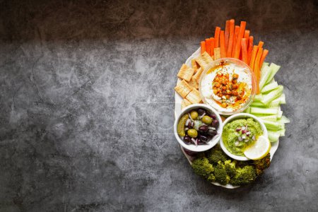 Photo for From above appetizing healthy hummus green sauce cut carrot cucumbers and green broccoli salted cucumbers decorated with sliced lemon in white bowls on table - Royalty Free Image
