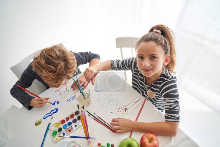 Photo for From above of content girl looking at camera while sitting near boy painting on paper at table with colorful paints - Royalty Free Image