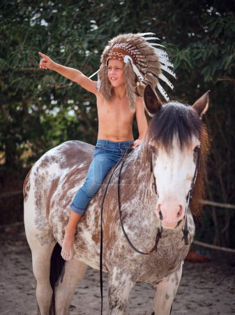 Foto de Niño montando a caballo en riendas mientras mira hacia el campo con árboles verdes y señalando de un lado a otro - Imagen libre de derechos