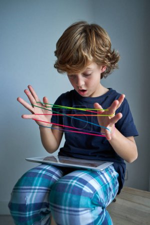 Photo for Shocked preteen child watching video on modern tablet while playing interesting cats cradle game on wooden stool in light room - Royalty Free Image