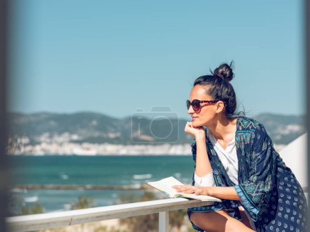 Téléchargez les photos : Vue latérale de magnifique femelle en vêtements d'été et lunettes de soleil assis avec livre sur la terrasse tout en admirant une vue imprenable sur la mer sur fond flou - en image libre de droit