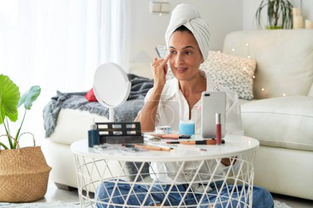 Photo for Smiling female influencer applying moisturizing facial cream on face skin in front of mirror while doing makeup and shooting video for beauty vlog at home in morning - Royalty Free Image