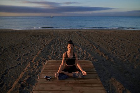 Téléchargez les photos : Corps complet de femme hispanique assise dans la posture Padmasana tout en méditant sur la promenade sur le rivage de sable pendant l'entraînement de yoga près de la mer - en image libre de droit