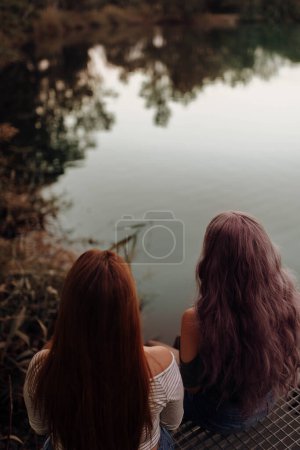 Téléchargez les photos : Forme ci-dessus vue arrière de jeunes femmes aux cheveux longs assis ensemble au-dessus de l'eau du lac tranquille - en image libre de droit