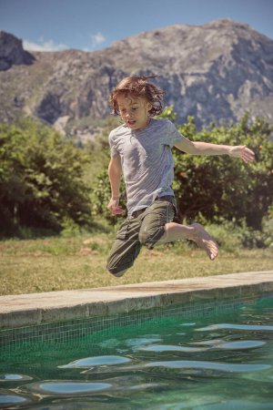 Téléchargez les photos : Corps entier d'adorable enfant excité en t-shirt et short sautant dans la piscine et regardant vers le bas contre la zone montagneuse floue en été - en image libre de droit