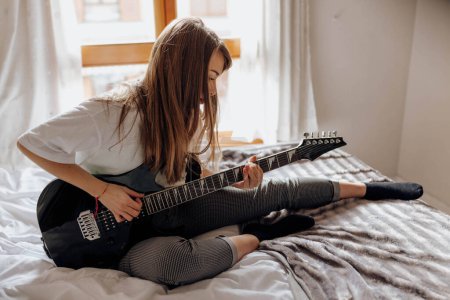 Téléchargez les photos : Vue latérale de Jeune femme brune jouant de la guitare sur le lit - en image libre de droit