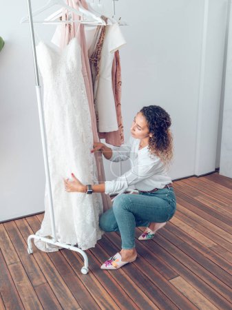 Photo for Side view of woman with curly hair in casual clothes squatting down and touching dress hanging on rack while choosing clothes in showroom - Royalty Free Image