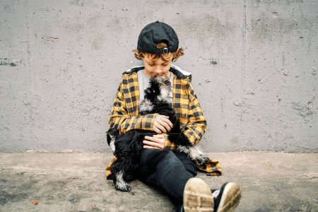 Téléchargez les photos : Mignon petit enfant en chemise à carreaux assis sur le sol en béton et jouer avec un petit chien tout en passant du temps ensemble - en image libre de droit