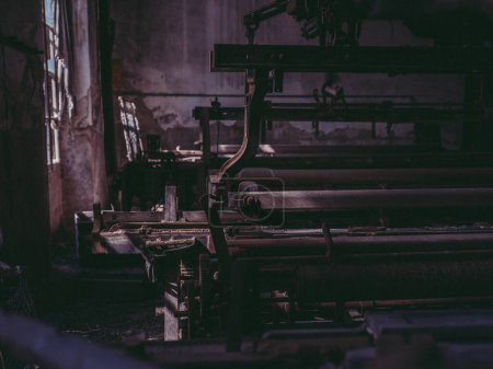 Foto de Antiguo telar de metal oxidado y máquina colocada cerca de la pared de piedra en la fábrica de hilo industrial abandonado oscuro - Imagen libre de derechos