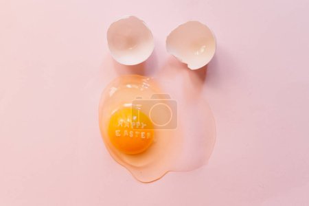 Foto de Huevo crudo roto de arriba con texto Feliz Pascua sobre yema amarilla y cáscaras de huevo sobre fondo rosa - Imagen libre de derechos