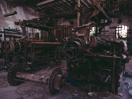 Téléchargez les photos : Métier à tisser en métal rouillé vieilli avec des engrenages et des équipements cassés placés sur le sol endommagé dans l'usine de fils industriels abandonnés grungy - en image libre de droit