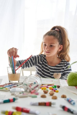 Photo for Concentrated girl choosing paintbrushes in glass with water while painting at messy table with scattered crayons and paints in light room - Royalty Free Image