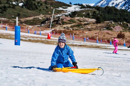 Foto de Niño optimista en ropa de abrigo y sombrero mirando a la cámara con sonrisa mientras se arrodilla en la nieve cerca del trineo de plástico en el día de fin de semana en la estación de esquí - Imagen libre de derechos
