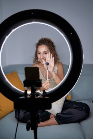 Photo for Full body of young female blogger showing professional brush for makeup while shooting video tutorial via cellphone in tripod using ring lamp while sitting on sofa - Royalty Free Image