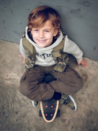 Photo for From above full body of adorable boy in stylish clothes sitting on skateboard leaning on wall on street while smiling and looking at camera - Royalty Free Image
