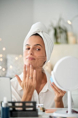 Foto de Encantadora mujer con turbante de toalla que aplica crema facial hidratante en la barbilla mirando el espejo colocado en la mesa - Imagen libre de derechos