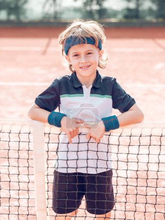 Téléchargez les photos : Petit garçon joyeux avec les cheveux blonds en uniforme et bandeau souriant et regardant la caméra tout en se tenant près du filet sur le terrain d'argile après l'entraînement de tennis le jour ensoleillé - en image libre de droit