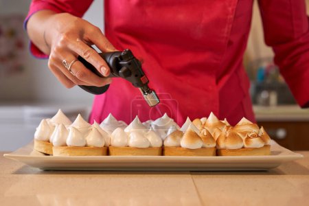 Foto de Crop cocinera femenina anónima con antorcha de gas preparando conjunto de merengue suizo en la mesa en la cocina - Imagen libre de derechos
