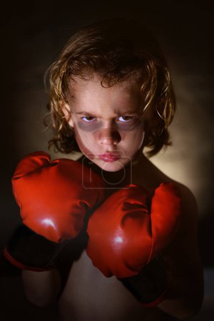 Foto de Niño serio con moretones y tiritas en la cara con guantes de boxeo rojos mirando a la cámara mientras está de pie sobre fondo oscuro - Imagen libre de derechos