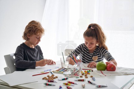 Téléchargez les photos : Enfants mignons peignant avec des peintures sur papier tout en étant assis à la table avec des fournitures d'art dispersés et des pommes dans la salle de lumière - en image libre de droit