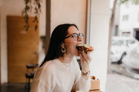 Téléchargez les photos : Vue latérale de jolie jeune femme en tenue élégante mordant sandwich savoureux tout en étant assis près de la fenêtre dans un café confortable - en image libre de droit