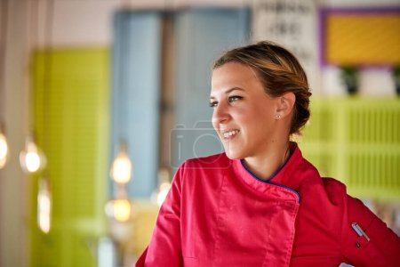 Téléchargez les photos : Portrait de cuisinière souriante portant une tenue professionnelle debout dans une pièce colorée sur fond flou tout en détournant les yeux - en image libre de droit