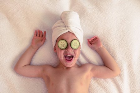 Foto de Vista superior del niño emocionado con toalla en la cabeza y rodajas de pepino en los ojos acostados sobre una manta blanca con los brazos levantados y riendo - Imagen libre de derechos