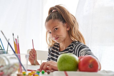 Téléchargez les photos : Adorable fille avec pinceau regardant la caméra tout en peignant à table avec des pommes et des bonbons dispersés tout en étant assis dans la salle de lumière - en image libre de droit