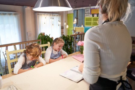 Photo for Back view of crop anonymous woman in apron standing at table and reading recipe for cheerful little sibling writing in notebooks before cooking in kitchen - Royalty Free Image
