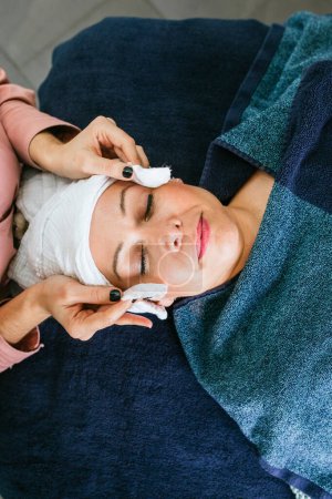 Photo for Top view of crop unrecognizable cosmetician using cotton pads and applying tonic on face of female customer lying on table in beauty salon during skincare procedure - Royalty Free Image
