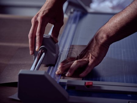 Artesano irreconocible que usa una máquina moderna para cortar trozos de papel mientras trabaja en la mesa en un taller de encuadernación