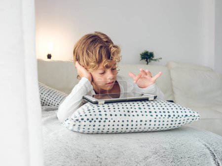Téléchargez les photos : Adorable garçon préadolescent couché sur un lit confortable navigation sur Internet sur tablette placée sur oreiller - en image libre de droit