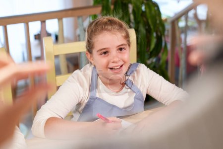 Photo for High angle of cheerful little schoolgirl with blond hair in casual clothes smiling while listening to crop anonymous teacher sitting at table with notebook and pen in hand - Royalty Free Image