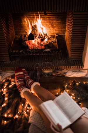 Téléchargez les photos : Crop femme en rouge chaussettes d'hiver assis devant la cheminée et lecture livre de papier tout en appréciant l'humeur du soir et de Noël - en image libre de droit