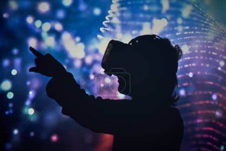 Foto de Vista lateral de un niño irreconocible con gafas VR modernas tocando la pantalla imaginaria mientras explora la realidad virtual en un cuarto oscuro con iluminación de neón brillante - Imagen libre de derechos
