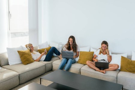 Téléchargez les photos : Amis féminins concentrés surfant sur les ordinateurs portables modernes et smartphone tout en étant assis sur un canapé confortable dans le salon lumineux avec table à la maison - en image libre de droit