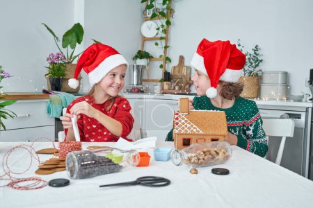 Téléchargez les photos : Frères et sœurs souriants en chapeaux de Père Noël se regardant tout en faisant maison de pain d'épice à table avec des biscuits pendant la préparation de Noël - en image libre de droit