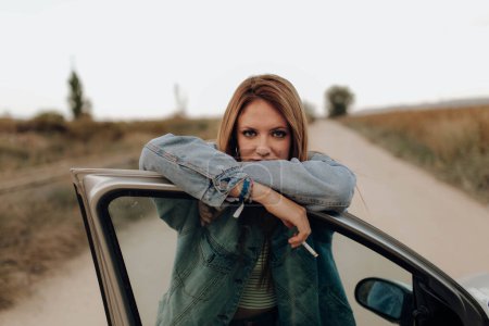 Téléchargez les photos : Femme confiante en jean veste debout avec cigarette à la porte de la voiture regardant caméra dans le pays - en image libre de droit