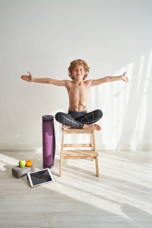 Foto de Cuerpo completo de adorable chico concentrado en pose de Loto con brazos extendidos mientras practica yoga en taburete de madera a la luz del día en la habitación - Imagen libre de derechos