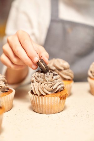 Téléchargez les photos : Cuisiner anonyme sans visage en plaçant un cookie sur des garnitures de chocolat de cupcake maison appétissant en mettant l'accent sélectif sur une table lumineuse - en image libre de droit